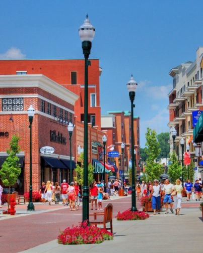 Sunny downtown shopping street with crowds strolling past brick buildings, lampposts, and flower planters under a blue sky.