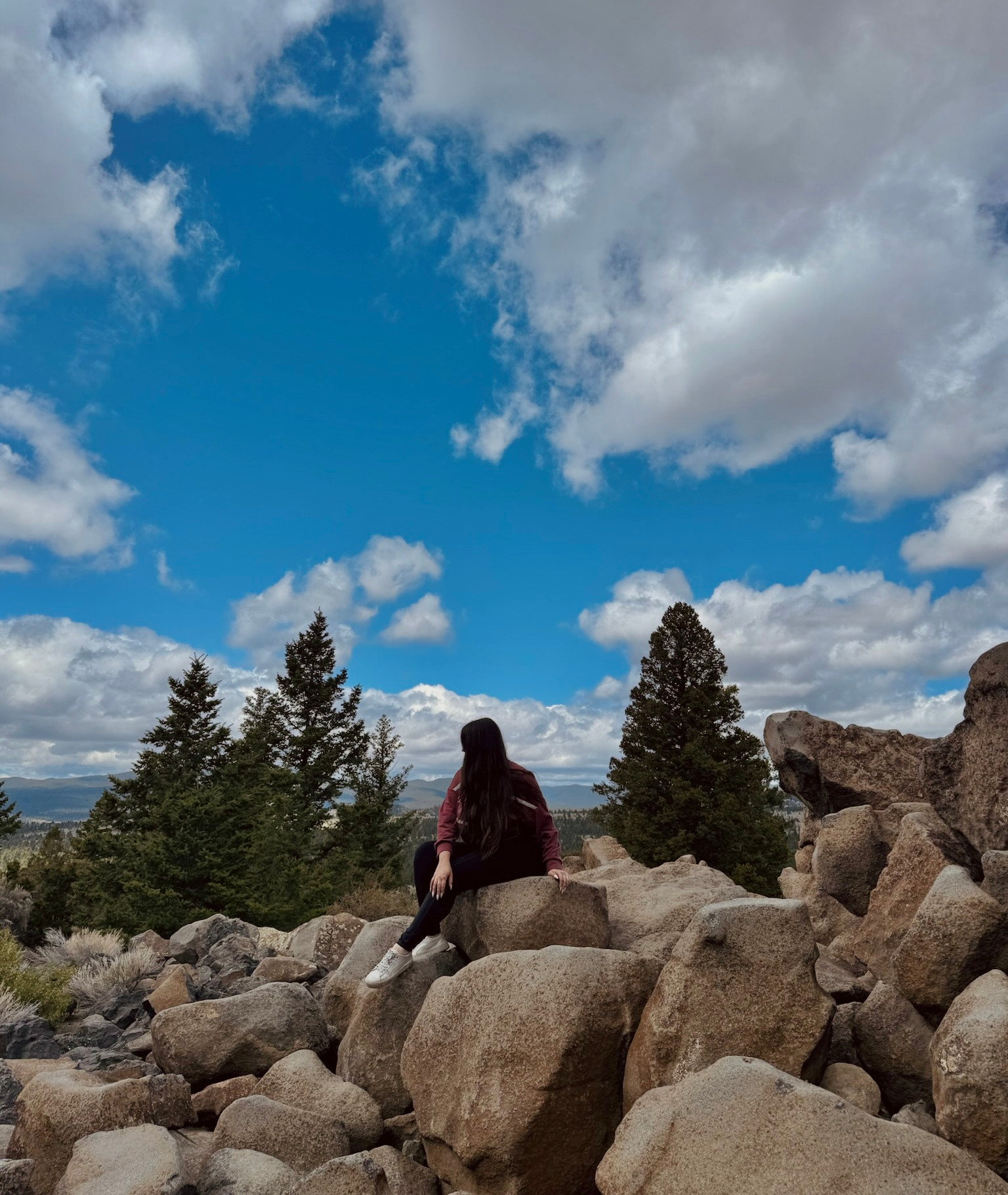 Woman in a maroon jacket sits on boulders beneath a dramatic blue sky, with pine trees and distant mountains behind her.