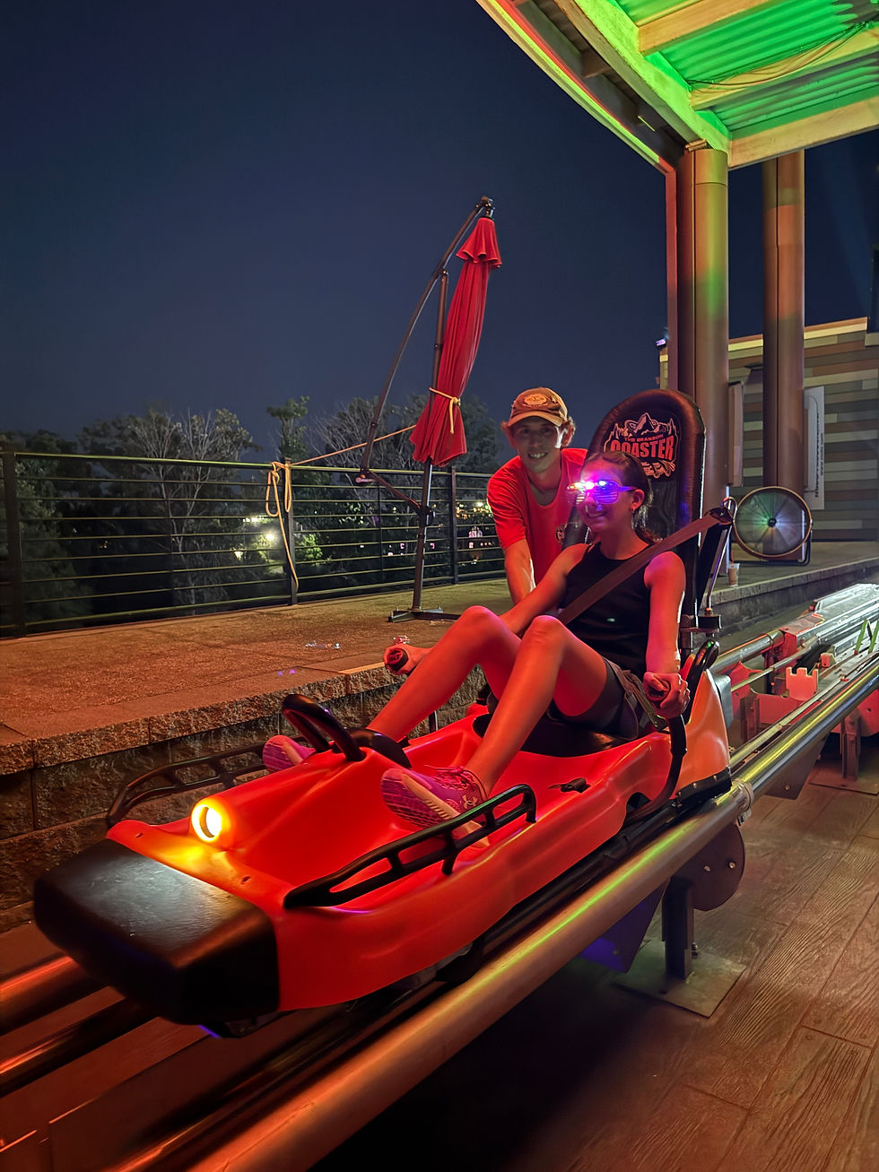 Two smiling riders on a red coaster car at night, with glowing goggles and a Branson Coaster seat back under a green-lit canopy.