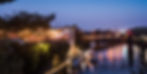 River at dusk with lit-up party venue on the left, a bridge in the background, and a boat docked below. Warm glowing lights create a festive mood.