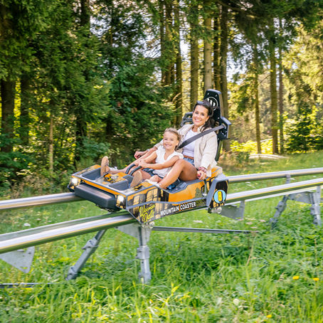 Child and woman happily riding a mountain coaster through a forest. The coaster is orange with "Mountain Coaster" text visible.