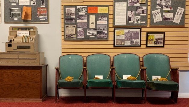 Museum exhibit of Jim Owen with old cash register, green chairs, and framed photos/text on a wood-paneled wall labeled OWEN BOAT LINE