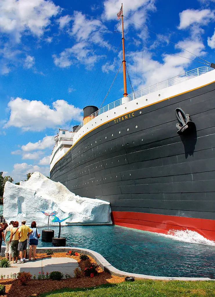 Titanic ship display beside a large iceberg, with tourists watching under a bright blue sky with clouds