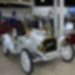 Vintage white Buick with gold trim in showroom. Giraffe statue behind car. Red wagon wheel visible. Bright, clean interior setting.