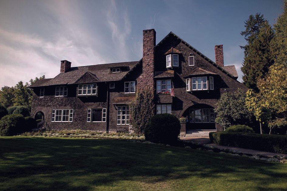 Large dark-shingled house with chimneys and many windows sits on a manicured lawn under a blue sky, framed by trees and shrubs