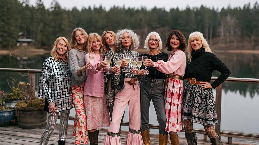 Seven stylish creative women toast with champagne on a lakeside deck, smiling in colorful retro outfits with forest and calm water behind. They are celebrating her successful business as a woman over 40.