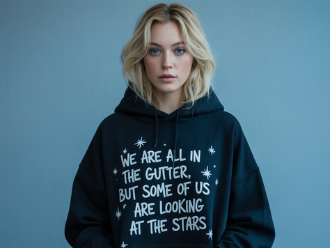 Blonde woman at The Yellow Studio in black print-on-demand hoodie with text: "We are all in the gutter, but some of us are looking at the stars." Blue background, neutral expression.