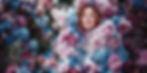 A woman with curly hair surrounded by pink and blue flowers and butterflies smiles softly, creating a whimsical and serene atmosphere. Image of Blume Bauer for the Yellow Studio.