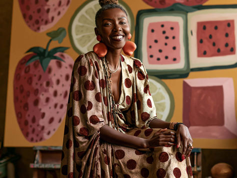 Smiling woman artist in a gold polka-dot dress sits in an art studio before a colorful fruit mural.