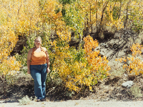Smiling woman, Blume Bauer, in an orange top and jeans stands among yellow and orange autumn trees on a sunny hillside. Camping at Rock Creek, near Bishop, California.