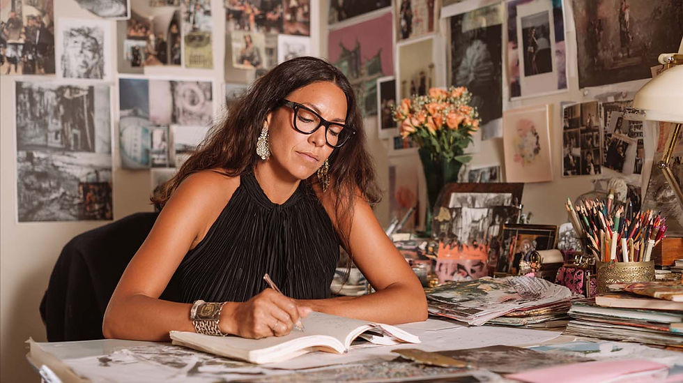 Woman with glasses writing in a notebook at a cluttered desk. Art-filled wall in the background. Flowers add a touch of color.
