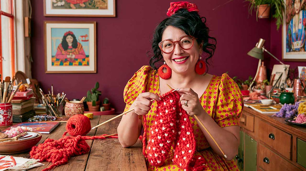 Smiling creative woman in glasses knits a red heart-pattern scarf at a wooden table in a colorful craft room.