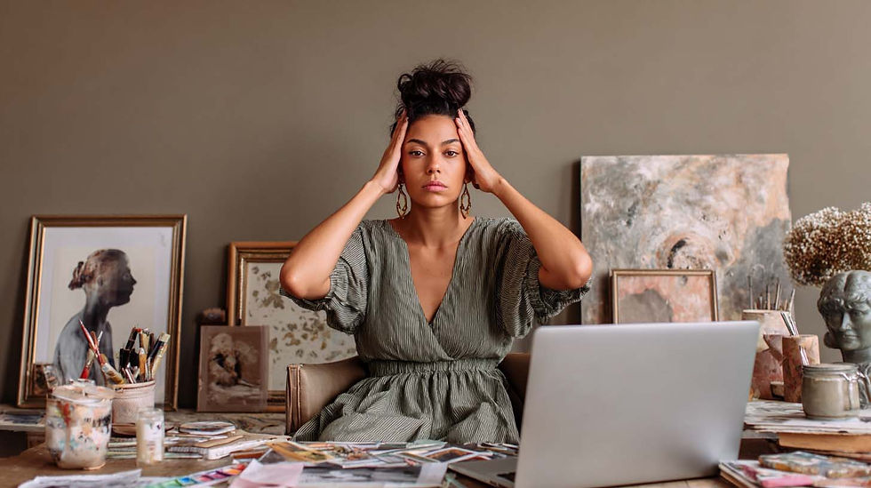 Woman in a green dress sits at a cluttered desk, holding her head, looking stressed. Art supplies and paintings surround her in a cozy room.