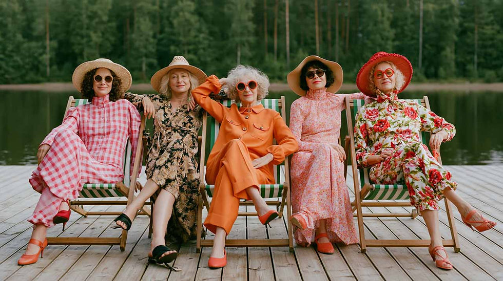 Five stylish creative women in colorful dresses and hats lounge on deck chairs by a lake, smiling in a relaxed summer scene. They are celebrating her successful business as a woman over 40.