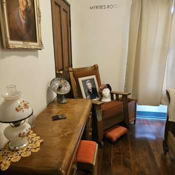 Vintage room with wooden chair, portrait and mannequin head by a table; wall text says Myrtie’s Room, softly lit and cozy. Interior room at the Western Hotel in Lancaster, California.