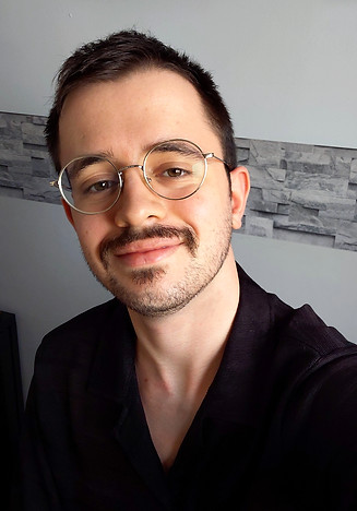 A young man with short dark hair, round glasses and facial hair. He is smiling, wearing a black shirt.