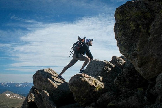 A hiker with a backpack climbs rugged boulders on a mountain peak under a blue sky. To reach his Vision. To fullfill his mission