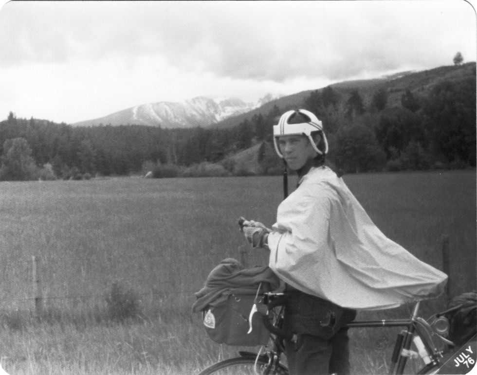 A young man straddles a bicycle wearing a bicycle helmet and a rain poncho. He is looking at the camera and holding something. There are clouds in the sky behind over mountains and trees.