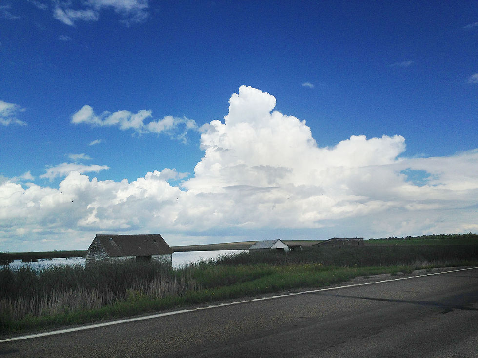 A landscape of a lake next to a road with an old, weathered building in the water. Big storm clouds are in the distance.
