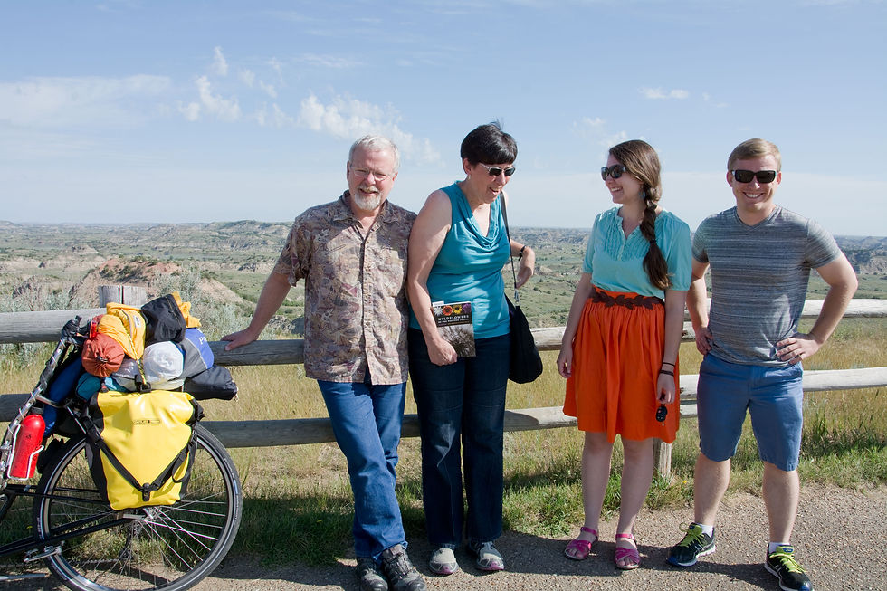 A landscape portrait of a man and woman with their grown daughter and her boyfriend standing in front of a wood fence next to a loaded recumbent bicycle. The background is the painted canyon.