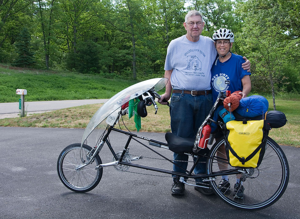 A man and woman stand behind a recumbent bicycle loaded with camping gear. The woman wears a helmet and the man has his arm around her shoulders.