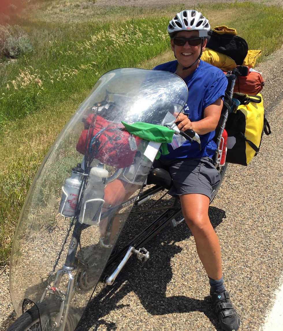 A woman wearing a white bicycle helmet and a blue shirt and shorts, sits on a loaded recumbent bicycle, which has a plastic fairing on its front. She is smiling, with grass in the background.