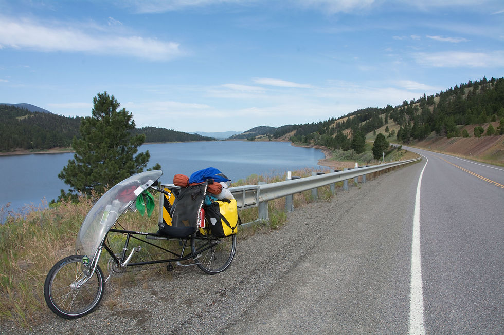 A landscape scene of a lake on the left, with grasses in the foreground next to a guardrail and a road between the lake and treed hills. In the foreground is a loaded recumbent bicycle leanig against the guardrail.