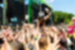 Crowd surfing at a sunny outdoor music festival, with hands raised before a giant green stage and screens.