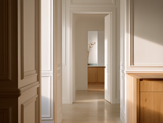 Corredor de vivienda reformada en Barcelona con molduras originales restauradas, suelo continuo de piedra clara y carpintería de roble natural, luz natural lateral