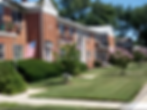 Exterior photograph of a red brick apartment building with manicured lawns. An American flag adorns the facade under a clear sky.