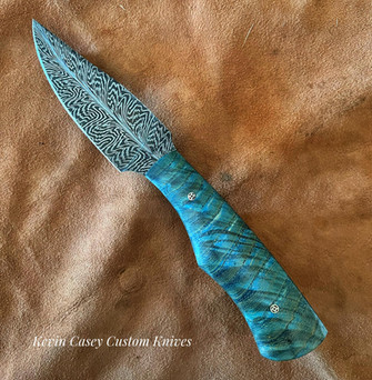 Knife by Kevin Casey. 4.25” Casey Feather Damascus blade. OAL 8.75”. Full tang construction. Dyed and stabilized Box Elder handle w/G10 liners. Mosaic pins. Tooled custom leather belt loop style sheath.