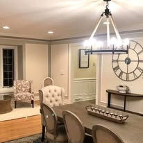 Interior dining room with table, chairs, chandelier, and wall clock in warm neutral colors, square aspect ratio.