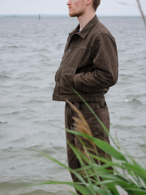man wears brown jacket and pants standing against a backdrop of gray blue water