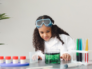 A young girl wearing safety goggles and a lab coat conducts a science experiment at a table with colorful test tubes and beakers filled with green liquid.
