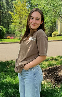 Woman with long dark hair wearing a Scott’s Coffee Co. shirt, smiling outdoors in a sunlit, natural setting.