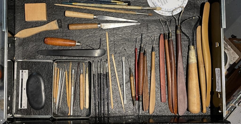 A drawer of sculpting tools on a black surface, including wooden handles, metal tips, and carving knives, arranged neatly.