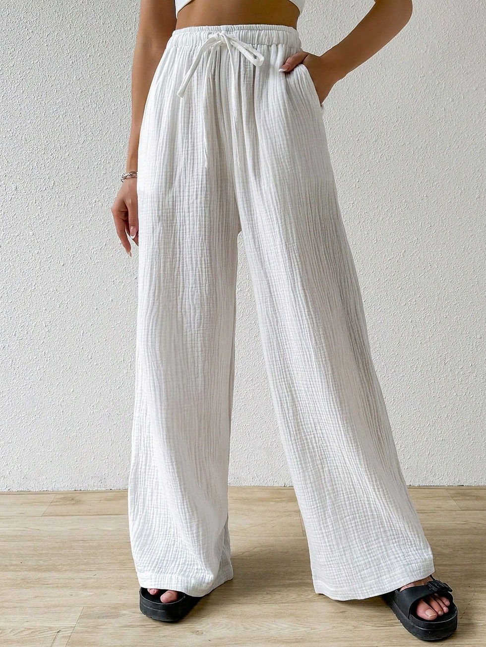White wide-leg pants made of textured fabric with a drawstring waist, worn by a woman standing against a white wall, paired with a cropped top and black sandals