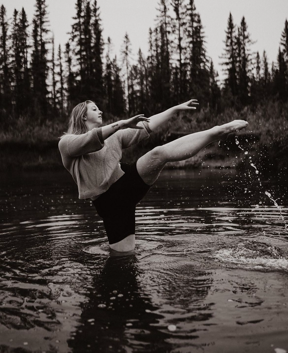 woman standing thigh deep in a lake the horizon is laced with trees, black and white photo, dancing in the water with one leg out in attitude flicking the water off her toes