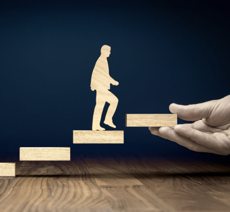A giant hand placing blocks in the air that a figurine is walking up: similarly to how a health and wellness coach helps build a path forward for coaching clients who specifically experience problems in decision making or seeing a way forward due to burnout, chronic stress, or chronic illness.