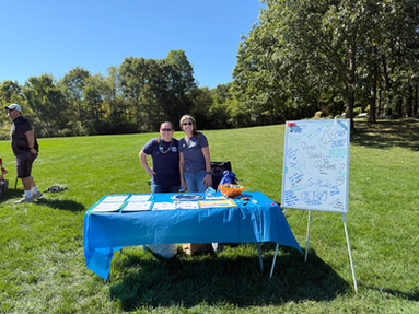 Volunteers stand behind an outdoor advocacy table with a whiteboard and flyers displayed.