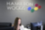 Woman working at a desk with computer monitors beneath the Hamilton Woods logo on a dark grey office wall