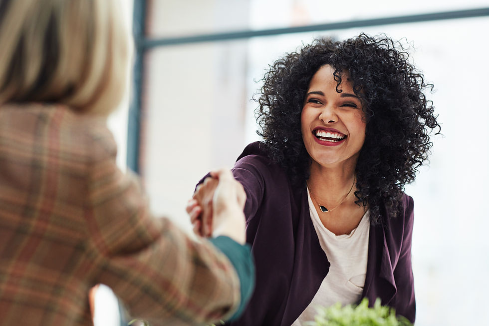 Smiling businesswoman with curly hair wearing a blazer, shaking hands with another person in a professional setting.