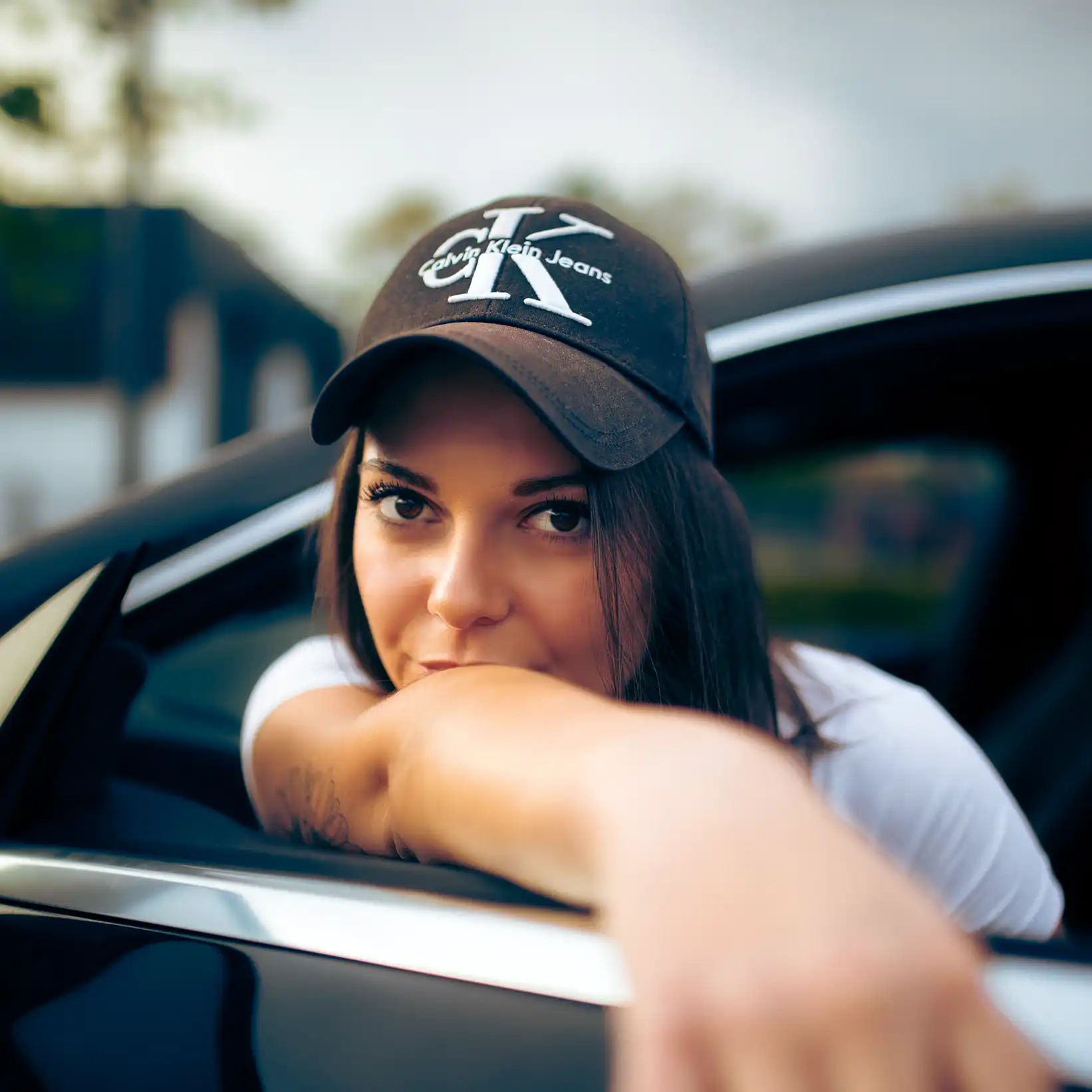 Portrait d'une femme acoudé à la portière d'une voiture noire. Elle porte une casquette noire avec un écriture blanche
