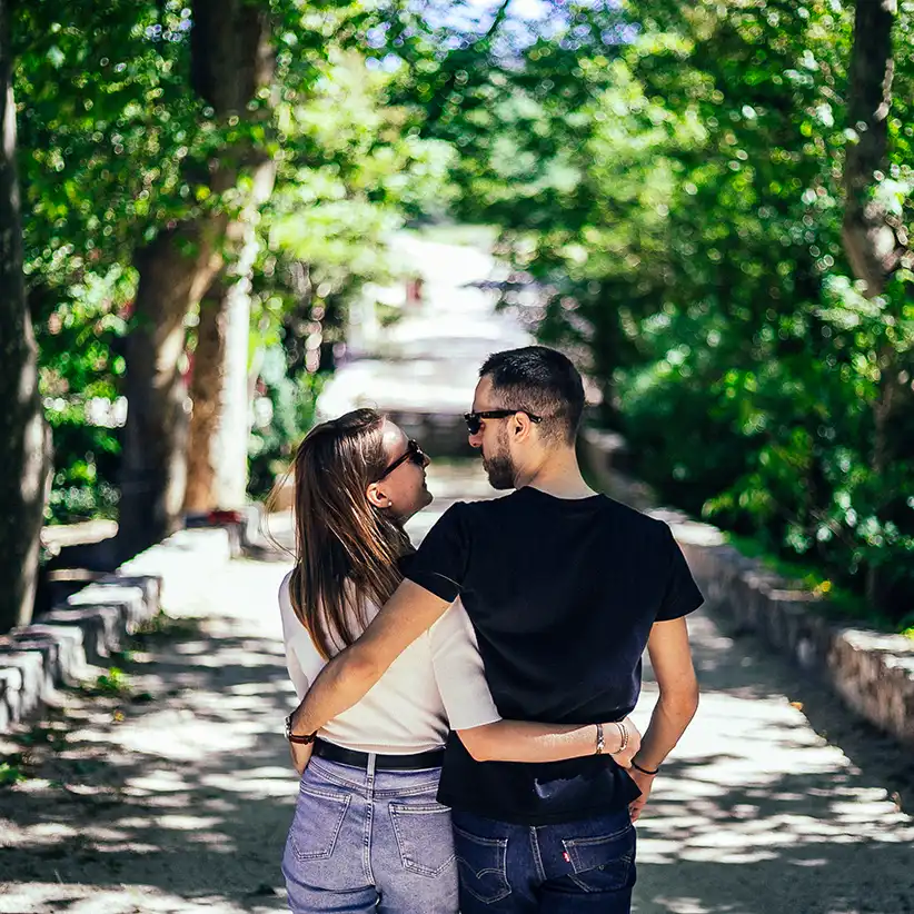 Portrait d'un couple marchant de dos sur un chemin entourée d'arbres. ils se regardent et portent des lunettes de soleil