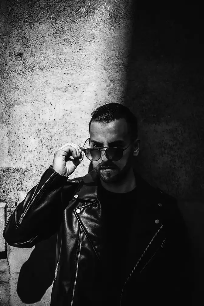 Portrait d'un homme en noir et blanc adossé à un mur et portant des lunettes noires. il porte également une veste en cuir
