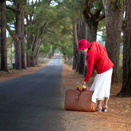 A woman bends to pick up her suitcase by a long country lane.
