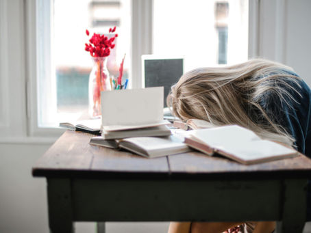 A woman with face buried on a desk covered in open books and a laptop.