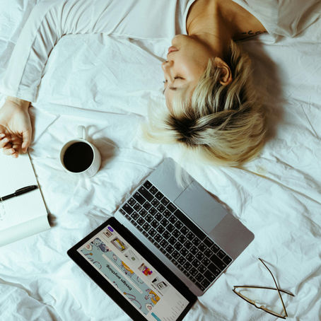A woman lying on her bed with a laptop, coffee, notebook and glasses