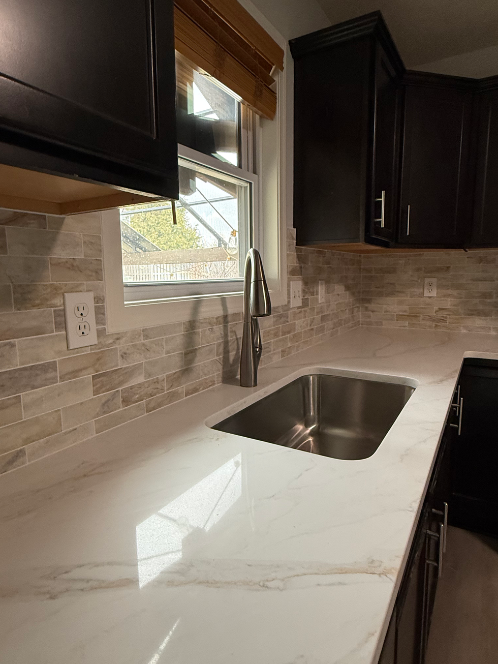 Kitchen sink with white quartz countertop, tile backsplash, and dark cabinets.