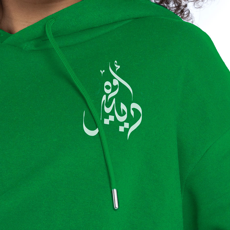 الصورة المصغرة: Christmas White Deer Written In Arabic Calligraphy front view on a green hoodie close up